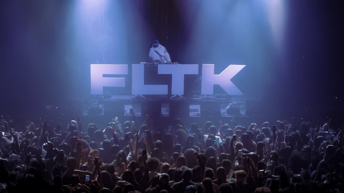 Flowtiko nos pratos por trás das letras gigantes FLTK iluminadas, diante de um público que filma com o telemóvel