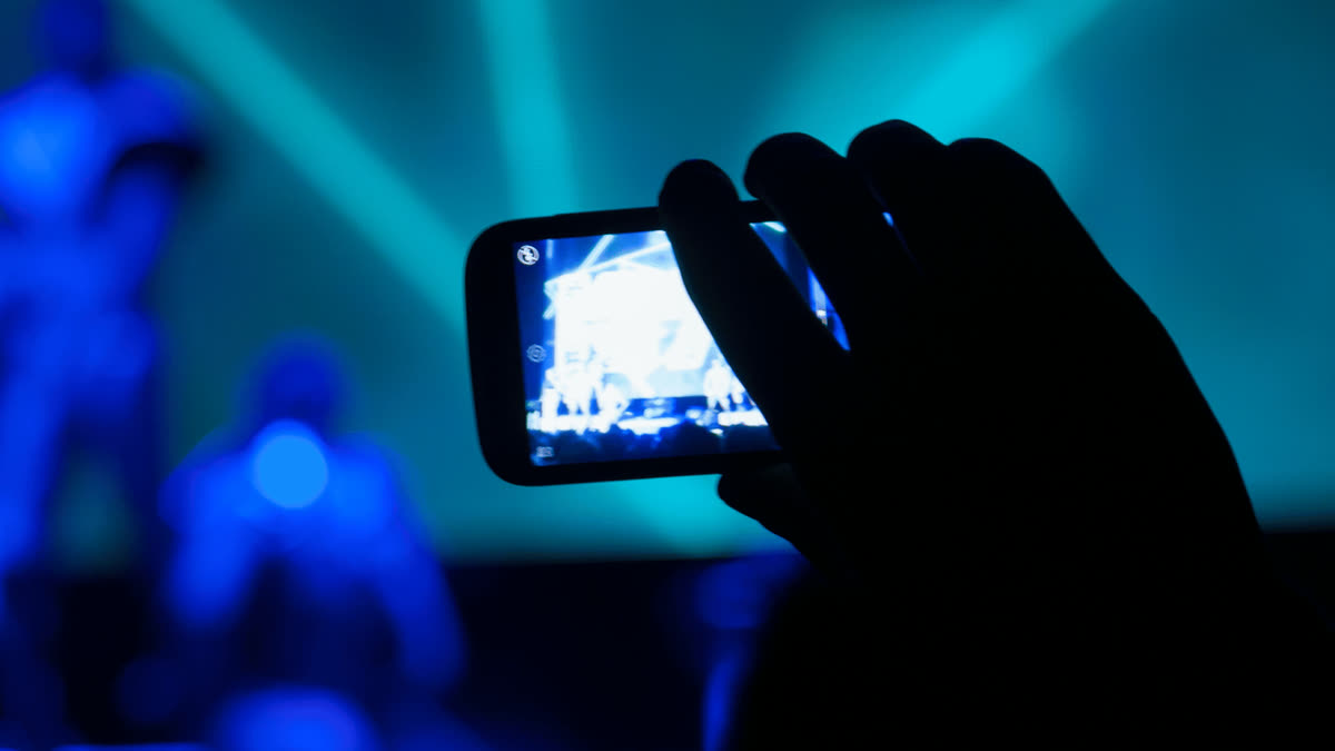 Silhouette einer Hand, die ein Konzert mit einem Smartphone filmt, unter türkisen und blauen Laserlichtern