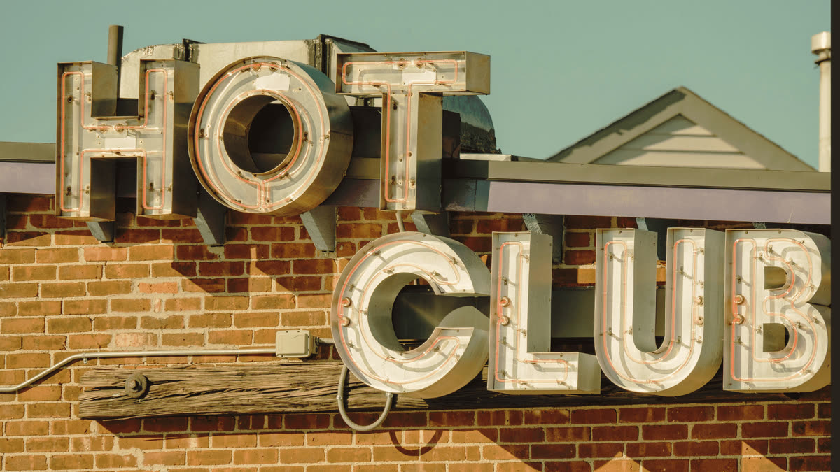 Neonschild „HOT CLUB“ an einer Backsteinmauer, vor blauem Himmel