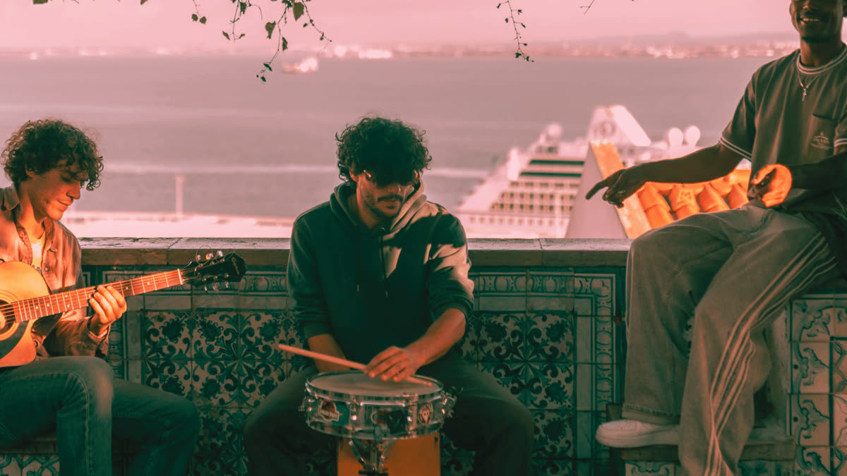 Three street musicians playing guitar and percussion facing the sea, under a tree